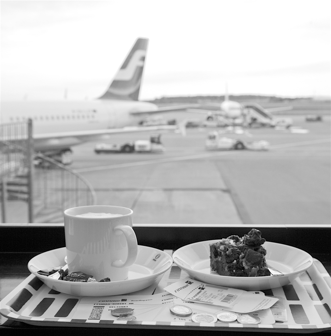 schwarz-weisses Tablett mit Cappucino, brownie, Bordkarte und Kleingeld am Flughafen